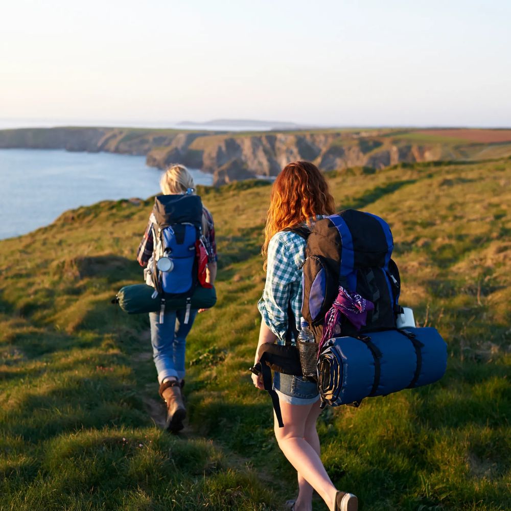 Two hikers with backpacks and camping gear walking along a grassy path near a body of water.
