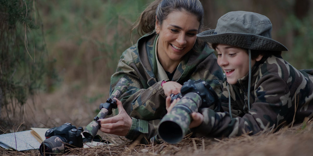 Woman and child in camouflage gear lying on the ground with binoculars in a forest setting